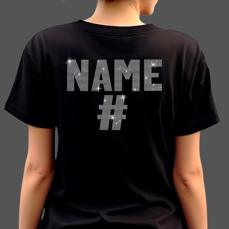 May include: A black short-sleeved t-shirt featuring the word NAME in large, silver-toned rhinestone lettering across the upper back, positioned above a silver-toned rhinestone hashtag symbol. The design is composed of small, reflective crystals.