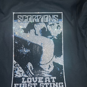 Peut inclure: T-shirt noir avec un motif en strass. Le motif comprend le nom du groupe "SCORPIONS" et la phrase "LOVE AT FIRST STING". L'image est un portrait d'une personne. Le t-shirt est &agrave; col rond.