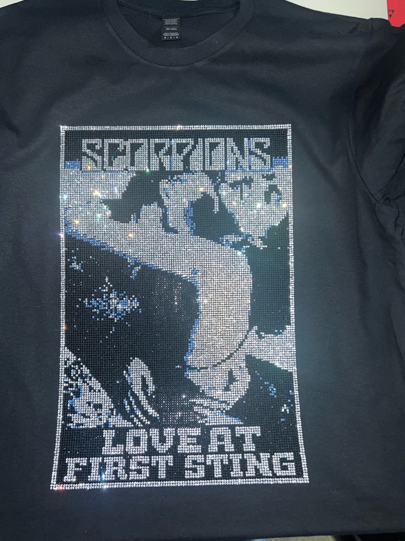 Peut inclure: T-shirt noir avec un motif en strass. Le motif comprend le nom du groupe "SCORPIONS" et la phrase "LOVE AT FIRST STING". L'image est un portrait d'une personne. Le t-shirt est &agrave; col rond.