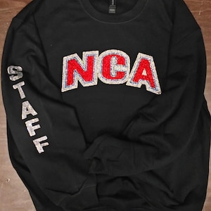 Op de afbeelding: Een zwart sweatshirt met ronde hals en de letters NCA in rode en zilveren pailletten op de borst. Het woord STAFF staat verticaal in zilveren pailletten op de rechtermouw. Het kledingstuk heeft lange mouwen en een geribbelde halslijn.