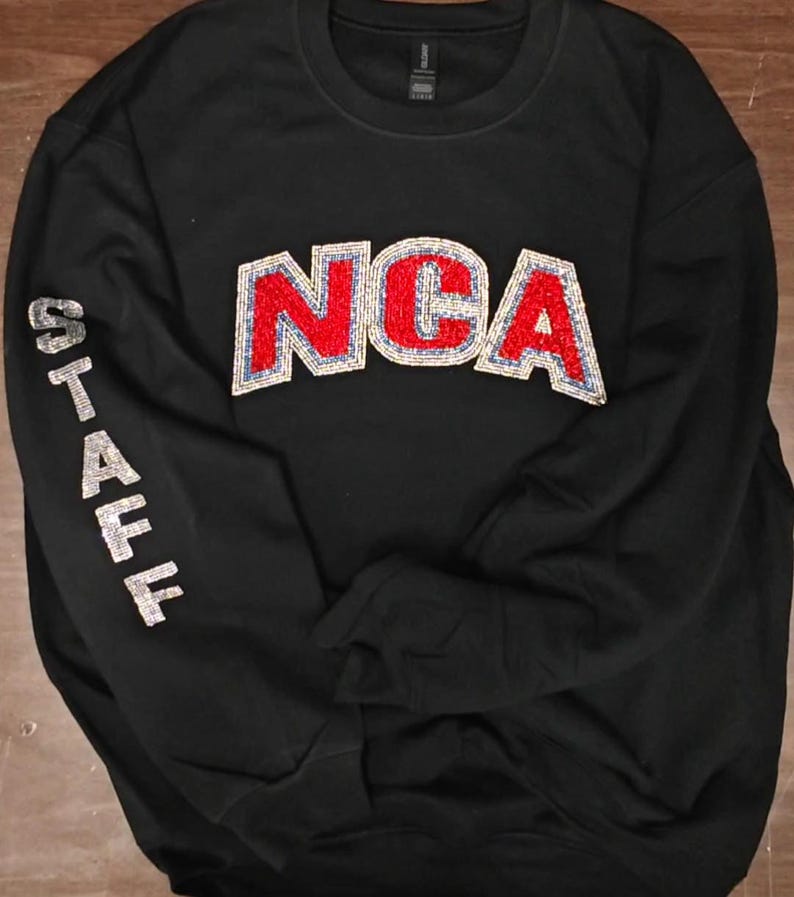 Op de afbeelding: Een zwart sweatshirt met ronde hals en de letters NCA in rode en zilveren pailletten op de borst. Het woord STAFF staat verticaal in zilveren pailletten op de rechtermouw. Het kledingstuk heeft lange mouwen en een geribbelde halslijn.