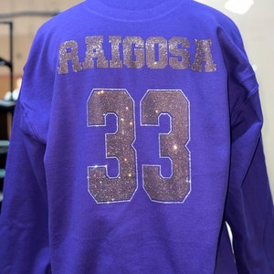 Op de afbeelding: Een paars sweatshirt met lange mouwen met de naam RAIGOSA en het nummer 33 op de achterkant. De tekst en het nummer zijn weergegeven in een bloklettertype gevuld met kleine, reflecterende, donkergekleurde strass-steentjes.