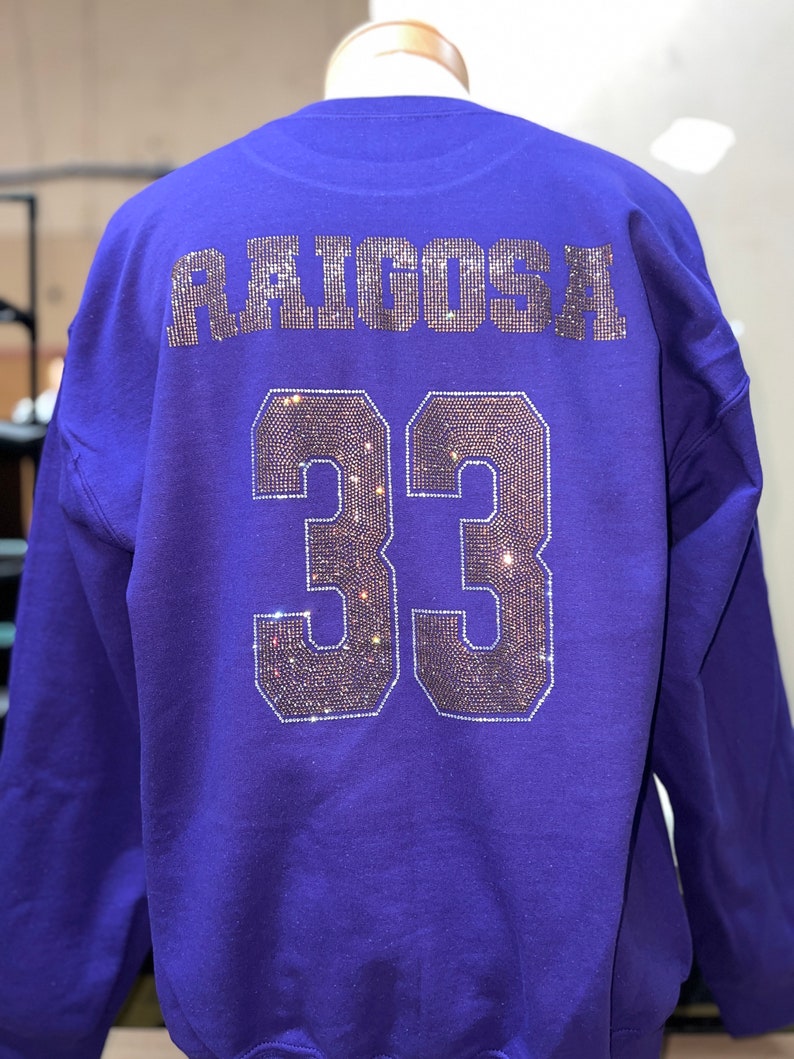 Op de afbeelding: Een paars sweatshirt met lange mouwen met de naam RAIGOSA en het nummer 33 op de achterkant. De tekst en het nummer zijn weergegeven in een bloklettertype gevuld met kleine, reflecterende, donkergekleurde strass-steentjes.