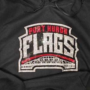Op de afbeelding: Een zwarte hoodie met een strass-afbeelding op de borst. Het ontwerp bevat de tekst PORT HURON in rode strass-steentjes boven het woord FLAGS in zilveren strass-steentjes, met daaronder een illustratie van een brug in zilveren en rode strass-steentjes.