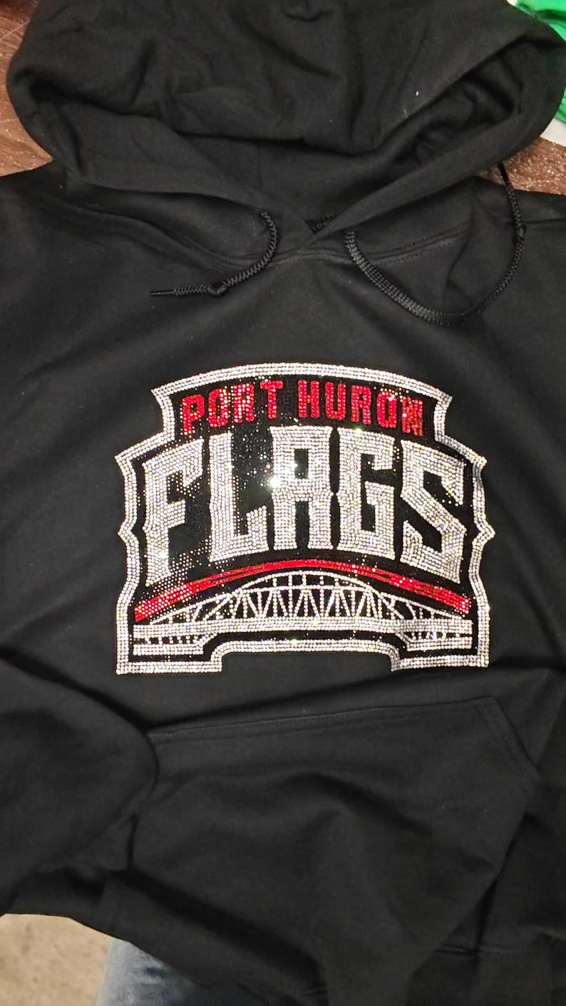 Op de afbeelding: Een zwarte hoodie met een strass-afbeelding op de borst. Het ontwerp bevat de tekst PORT HURON in rode strass-steentjes boven het woord FLAGS in zilveren strass-steentjes, met daaronder een illustratie van een brug in zilveren en rode strass-steentjes.