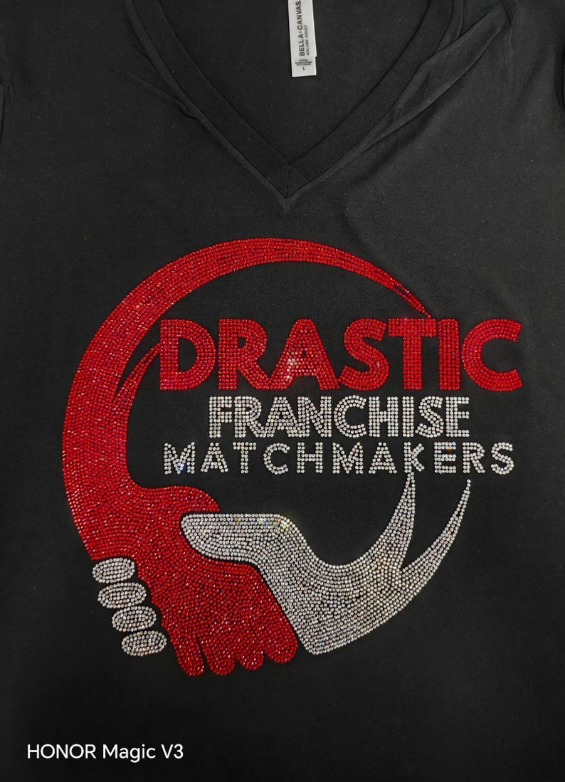 Peut inclure: T-shirt noir &agrave; col en V avec un motif en strass. Le motif pr&eacute;sente le mot "DRASTIC" en strass rouges et les mots "FRANCHISE MATCHMAKERS" en strass argent&eacute;s, ainsi qu'un graphisme rouge et argent.