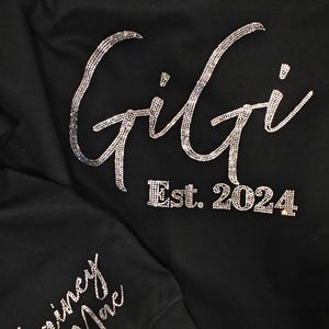 Op de afbeelding: Een zwart sweatshirt met lange mouwen voorzien van het woord Gigi en Est. 2024 in zilveren strass-steentjes op de borst. De mouw toont de naam Lainey Mae in zilveren strass-steentjes met een klein rood strass-hartje.