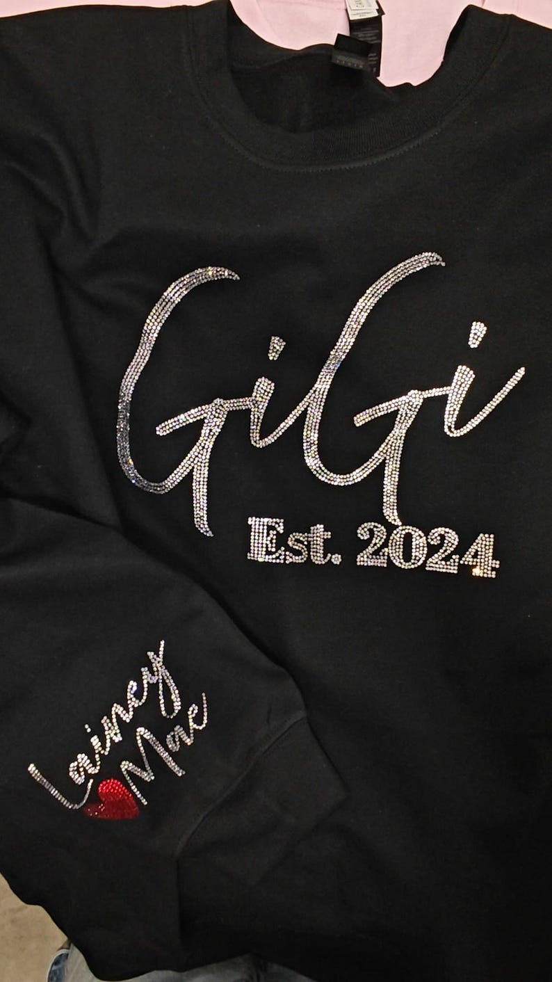 Op de afbeelding: Een zwart sweatshirt met lange mouwen voorzien van het woord Gigi en Est. 2024 in zilveren strass-steentjes op de borst. De mouw toont de naam Lainey Mae in zilveren strass-steentjes met een klein rood strass-hartje.