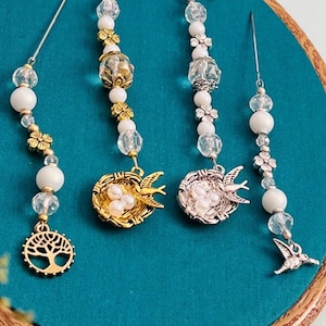 May include: Four decorative pin accessories with white beads, clear crystals, and gold or silver-toned charms. Charms include a tree of life, bird nests, and birds. The pins are displayed on a teal fabric within a wooden embroidery hoop.