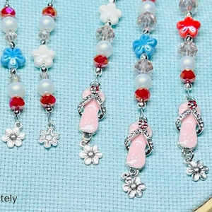 May include: A collection of beaded mask chains with various charms. The chains feature red, white, and clear beads, along with flower and star-shaped charms. Some chains have pink flip-flop charms. The text "*sold separately" is at the bottom.