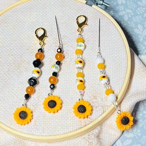 May include: Four needle minders featuring sunflower charms and assorted beads in yellow, black, and white. Each needle minder includes a gold-toned lobster clasp and a long metal needle attached to the beaded strand. The text sold separately is visible.