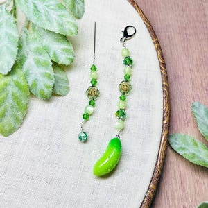 May include: Two beaded charms with green and gold accents. One charm has a green pickle pendant, the other has a needle. The charms are attached to a black clasp and a needle. The beads are various shades of green and white.