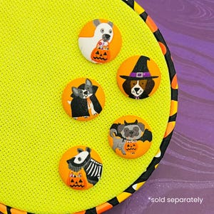 May include: A collection of five orange and black Halloween-themed buttons featuring dogs in costumes. The dogs are dressed as a vampire, witch, and holding pumpkins. The buttons are on a yellow background.