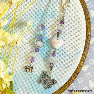 May include: Two beaded charms with butterfly and heart pendants. One charm has a silver butterfly pendant and a pink heart-shaped bead. The other charm has a silver heart pendant and a small butterfly charm. The charms are adorned with purple and clear beads.