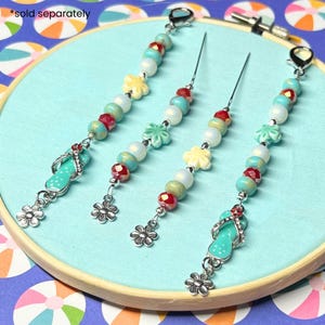 May include: Four beaded charm bracelets with turquoise, white, and red beads, and flower and flip-flop charms. The bracelets are displayed on a light blue fabric stretched over a wooden embroidery hoop.
