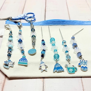 May include: A collection of seven winter-themed stitch markers and pins featuring blue and white beads, snowflake accents, and enamel charms including a penguin, a beanie, a sweater, a mug, and a mitten, arranged on a light fabric pouch.