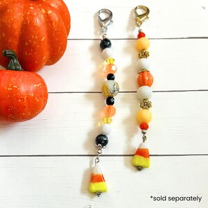 May include: Two beaded Halloween-themed charms with lobster claw clasps. One charm features black, white, orange, and yellow beads, and a candy corn charm. The other has orange, white, and yellow beads, and a candy corn charm. Two orange pumpkins are in the background.