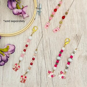 May include: Several beaded charms with gold-colored clasps and decorative charms. The charms feature pink, white, and red beads, along with small decorative charms. Some charms have a gift box design, while others have a teddy bear or pig design.