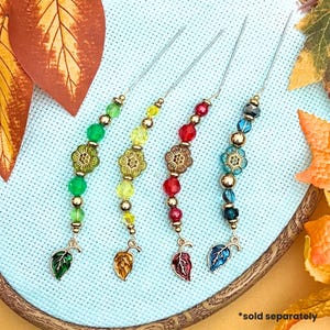 May include: Four colorful needle minders with beaded accents and leaf charms. Each needle minder features a different color scheme: green, yellow, red, and blue. The needle minders are attached to a light blue fabric background.