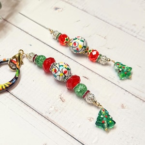 May include: Two decorative beaded ornaments featuring white spherical beads painted with colorful Christmas light patterns, accented by red and green faceted beads, silver spacers, and a small green glass Christmas tree charm at the end.