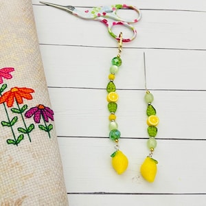 May include: A pair of beaded scissor fobs with lemon and leaf charms. The fobs are attached to floral-patterned scissors. A needle is also attached to one of the fobs. A floral embroidered fabric is in the background.