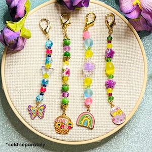 May include: Four beaded keychains with gold clasps. Each keychain features colorful beads, charms, and a unique pendant: a butterfly, a basket, a rainbow, and an Easter egg. The keychains are displayed on a white circular surface.