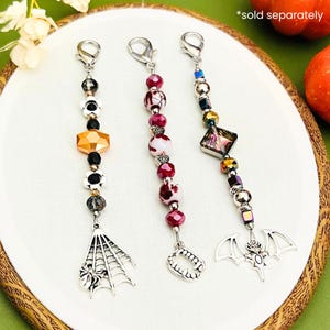 May include: Three Halloween-themed beaded charms with silver-tone lobster clasps. One charm features a spider web, another has a set of fangs, and the third has a bat. The beads are in various colors, including orange, black, red, and iridescent.