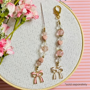 May include: Two beaded charms with pink heart and bow accents. One charm has a needle attached. The charms are gold and pink, with clear beads. The charms are on a white background with pink flowers.