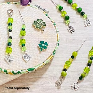 May include: Close-up of St. Patrick's Day themed accessories. The image features green and silver beaded charms with clover pendants, along with two green enamel clover pins. The accessories are displayed on a white surface with a floral patterned border.