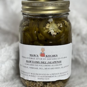 May include: A glass jar filled with sliced green jalapeños in a brine. The jar has a gold lid and a white label with the text "MAW'S KITCHEN" and "MAW'S OMG DILL JALAPENOS". The label lists ingredients and allergen information.