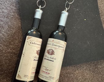 May include: A pair of novelty earrings featuring miniature wine bottle pendants. The black bottles have silver-colored tops and cream-colored labels with text. The earrings hang from silver-colored fish hook ear wires.
