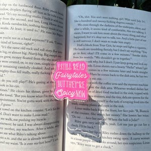 Puede incluir: Una pegatina rectangular de color rosa brillante con esquinas redondeadas que presenta el texto blanco I STILL READ Fairytales BUT THEY'RE Spicy NOW, colocada sobre las páginas abiertas de un libro con texto negro sobre papel blanco.
