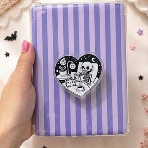May include: A rectangular tablet case featuring vertical purple and light purple stripes. A heart-shaped grip is attached to the center, displaying a black and white illustration of a skeleton reading in bed with a cat and a cup of coffee.