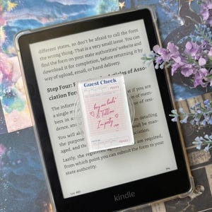Puede incluir: Un pisapapeles rectangular de acrílico transparente con un diseño de nota de restaurante impreso y el texto 'buy me books & tell me I'm pretty' en cursiva rosa, sobre un lector electrónico Kindle que muestra texto sobre formularios.