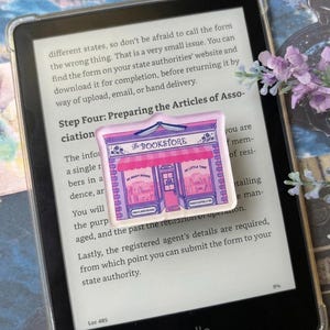 Op de afbeelding: Een rechthoekige acrylspeld met een roze en paarse illustratie van een boekwinkel met de tekst The BOOKSTORE, SO MANY BOOKS, SO LITTLE TIME en COFFEE & TEA, geplaatst op het scherm van een zwarte Kindle e-reader.