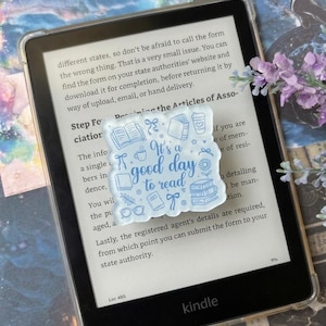 Op de afbeelding: Een vierkante, doorschijnende acrylspeld met blauwe lijntekeningen van boeken, een koffiekopje, een bril en een bloem, met de tekst "It's a good day to read" in het midden, rustend op het scherm van een zwart e-reader-apparaat.