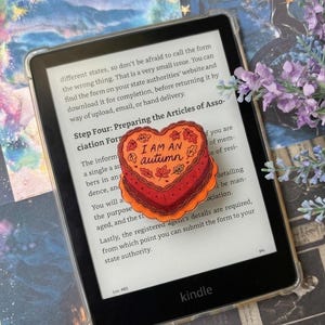 Op de afbeelding: Een hartvormige oranje en rode geborduurde patch met de tekst I AM AN AUTUMN omringd door kleine bladmotieven. De patch is geplaatst op het scherm van een zwarte e-reader die tekst over statutenformulieren weergeeft.