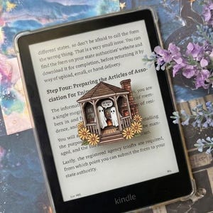 Può includere: Un lettore e-book nero che mostra del testo, con un segnalibro o magnete in legno a forma di gazebo contenente due figure di fantasmi bianchi, un camino in mattoni e fiori gialli alla base, con il testo SO SOON I'LL BE.