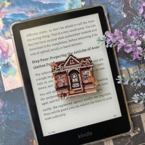 Op de afbeelding: Een zwarte Kindle e-reader met een doorzichtige beschermhoes toont tekst op het scherm. Een kleine, rechthoekige houten speld in de vorm van een boekhandel met herfstbladeren en pompoenen ligt op het scherm en bedekt een deel van de tekst.