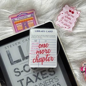 May include: Three acrylic pins featuring a bookstore storefront, a pink sign with cherries and the text Buy me books & tell me I'm pretty, and a library card design with the text one more chapter, resting on a book titled S-CORP & TAXES.