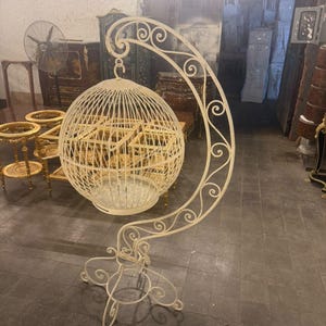 May include: A cream-colored, spherical metal birdcage suspended from a tall, curved, scroll-work stand. The cage features vertical wire bars and a circular base, while the stand has a decorative, ornate metal frame with a stable four-legged base.