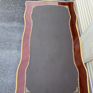 May include: A rectangular desk with a dark brown leather top and a decorative border. The border is made of reddish-brown wood with a gold trim. The desk has a unique, curved shape.