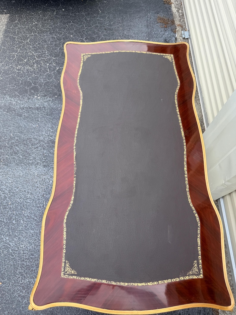 May include: A rectangular desk with a dark brown leather top and a decorative border. The border is made of reddish-brown wood with a gold trim. The desk has a unique, curved shape.