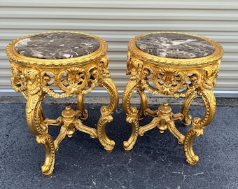 May include: Two ornate, gold-colored side tables with marble tops. The tables have intricately carved details and curved legs. The marble tops feature a brown and gray pattern. The tables are likely antique or vintage furniture.