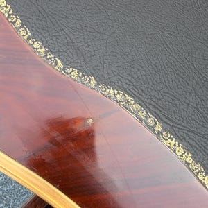 May include: Detail of a wooden table with a dark, polished surface. The table features a black leather top with a gold floral trim. The wood has a deep reddish-brown colour.
