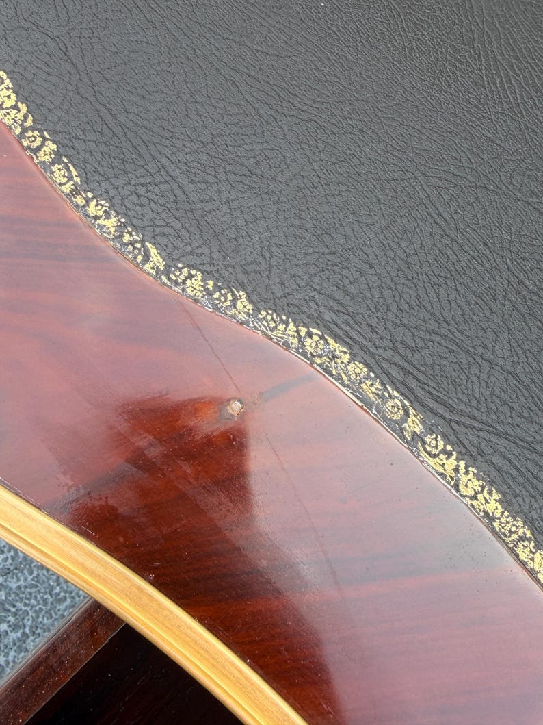 May include: Detail of a wooden table with a dark, polished surface. The table features a black leather top with a gold floral trim. The wood has a deep reddish-brown colour.