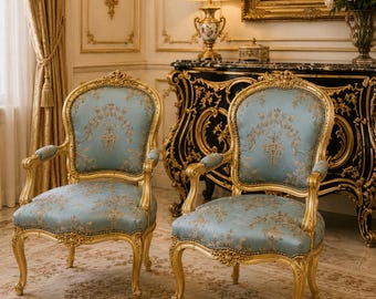 May include: A pair of armchairs featuring gold-painted wooden frames with intricate carved details and light blue upholstery adorned with a gold floral pattern. The chairs have curved legs, padded armrests, and high, rounded backrests.