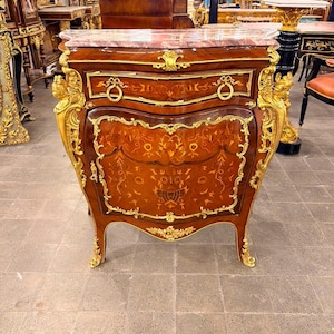 May include: A mahogany commode featuring a pink marble top, intricate floral marquetry on the front door, and ornate gold-toned metal mounts. The piece includes one drawer with two circular pulls and a single door, supported by four curved legs.