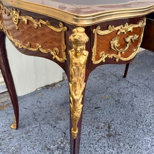 May include: An antique desk with a dark brown leather top and ornate gold accents. The desk features a curved design with detailed carvings and a rich, dark wood finish. The legs are slender and tapered, with gold detailing.