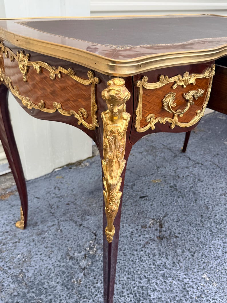 May include: An antique desk with a dark brown leather top and ornate gold accents. The desk features a curved design with detailed carvings and a rich, dark wood finish. The legs are slender and tapered, with gold detailing.
